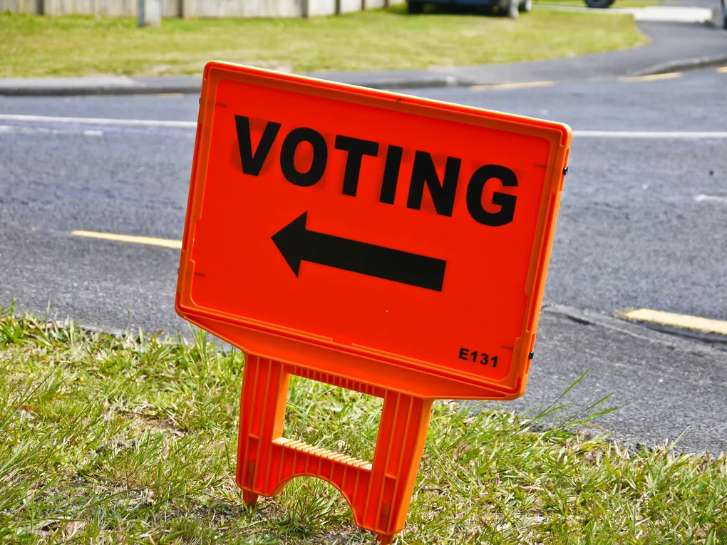 An orange sign outside a New Zealand polling place reading "VOTING", with an arrow pointing to the left